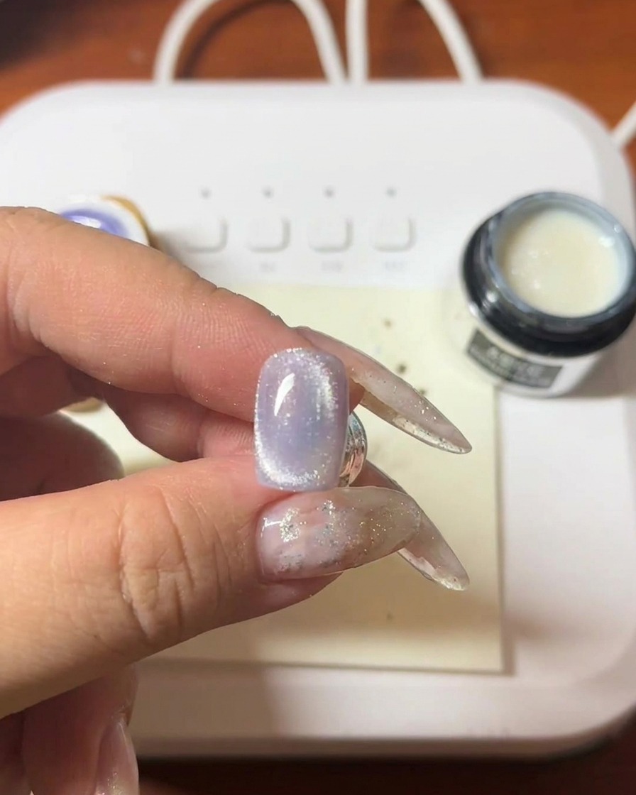 Zbliżenie na dłoń trzymającą różdżkę magnetyczną nad fioletowym paznokciem, widać formującą się jasną smugę cat-eye.