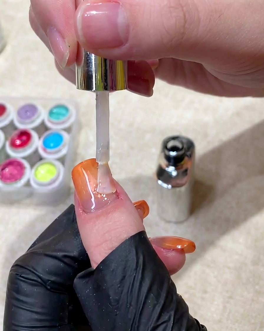 Pędzelek nakładający przezroczysty, błyszczący top coat na gotowy pomarańczowy paznokieć z gradientem i efektem cat-eye.