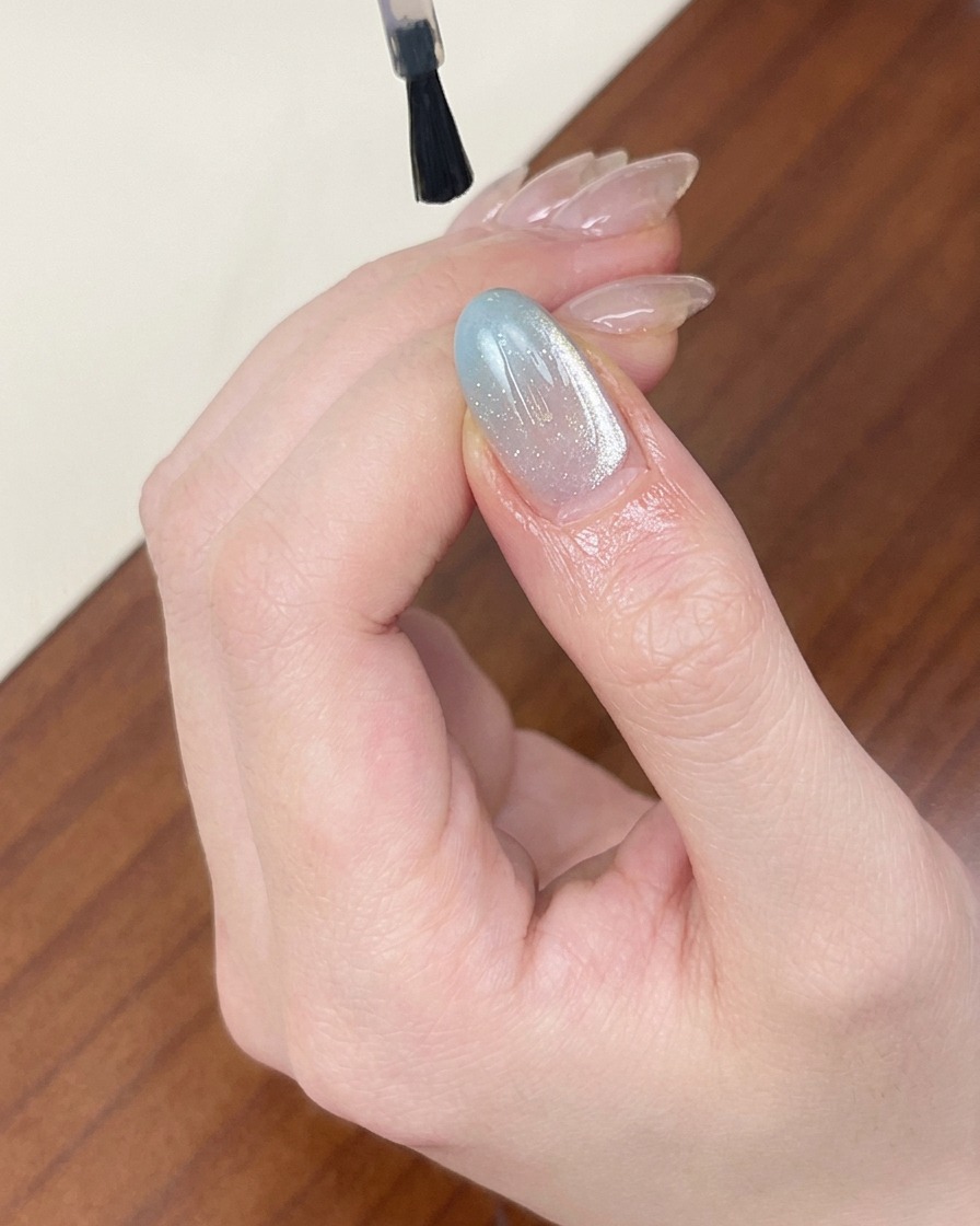 Pędzelek nakładający przezroczysty, błyszczący top coat na jasnoniebieski, mieniący się paznokieć z efektem cat-eye.
