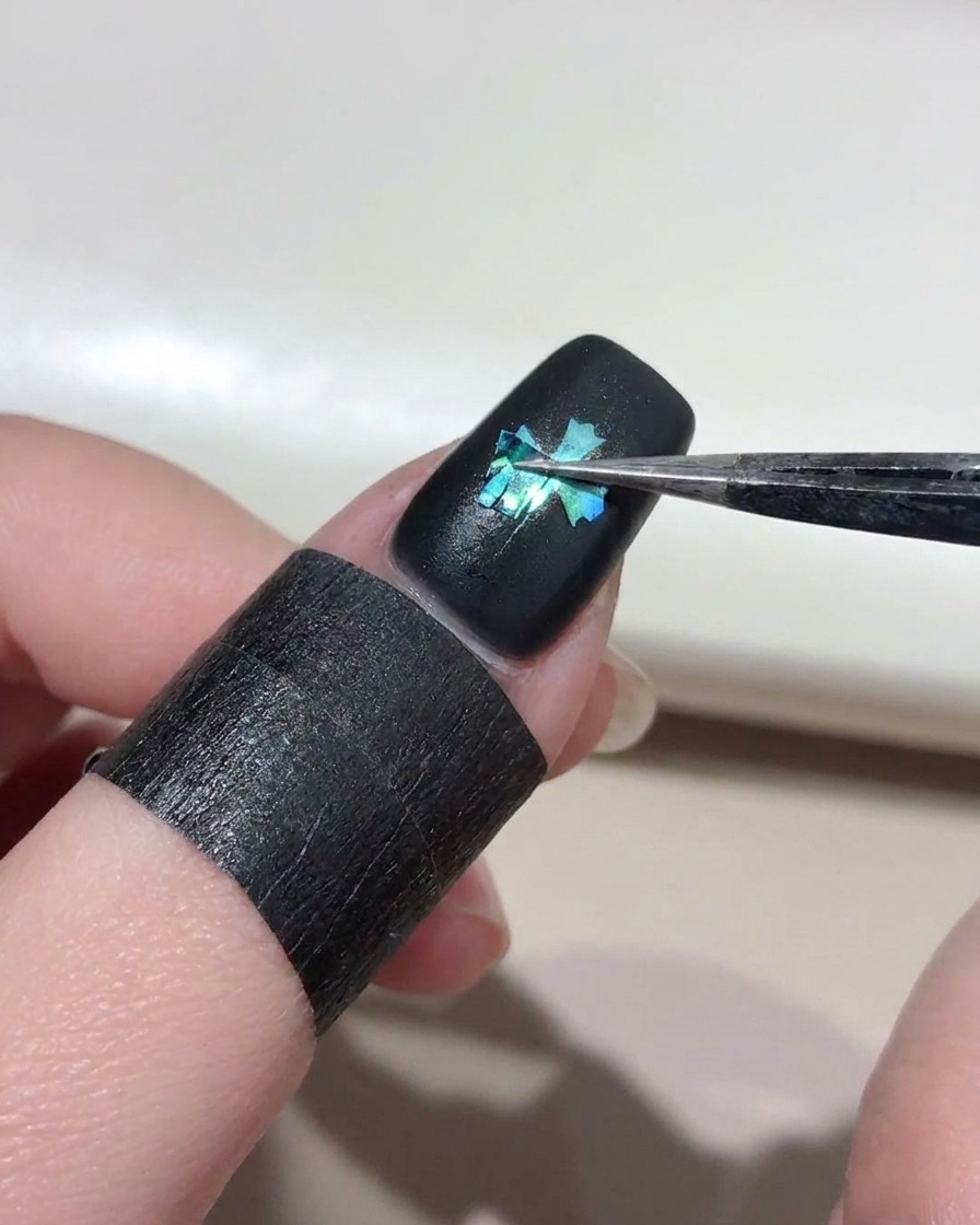Pęseta ostrożnie zdejmująca szablon krzyża z czarnego paznokcia, odsłaniając jasnoniebieski wzór krzyża cat-eye pod spodem.