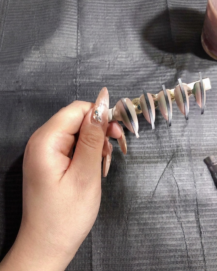 Pistolet airbrush rozpylający cienką warstwę białego żelu na sztuczne paznokcie zamontowane na stojaku.
