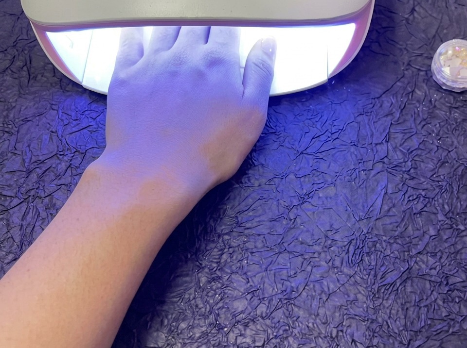 Dłoń spoczywająca we wnętrzu świecącej lampy UV/LED do paznokci, umieszczonej na fioletowej powierzchni o wyraźnej teksturze.
