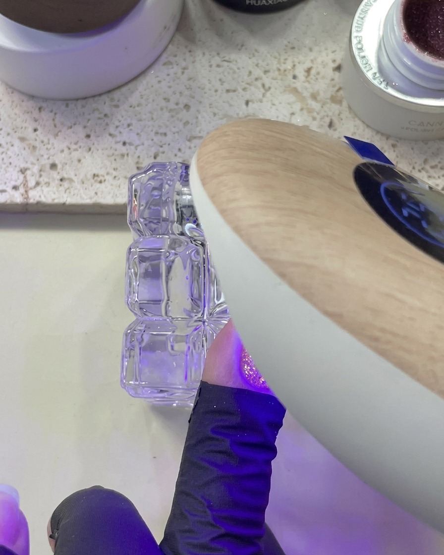 Dłoń umieszczona pod lampą UV/LED podczas utwardzania paznokcia z różowym brokatem.