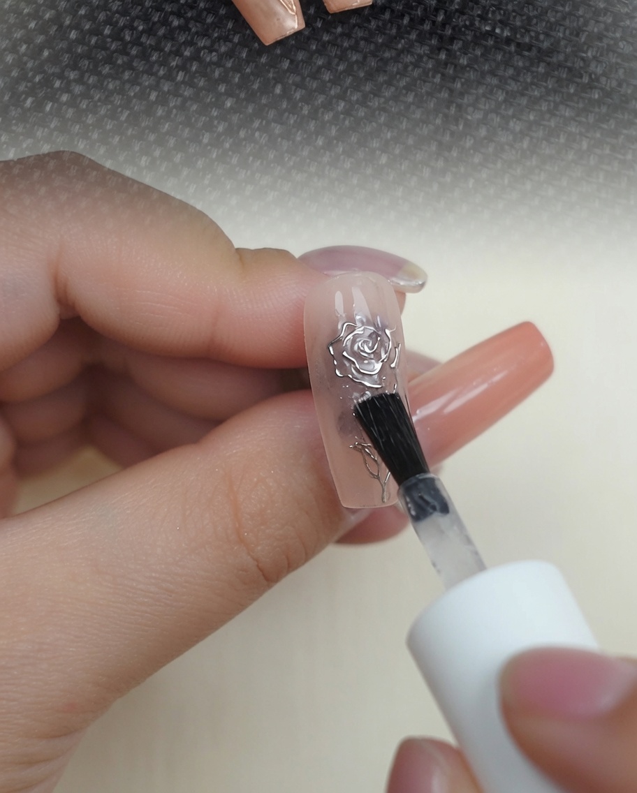 Pędzelek nakładający przezroczysty, lśniący top coat na metaliczną różę 3D na paznokciu w kolorze nude.
