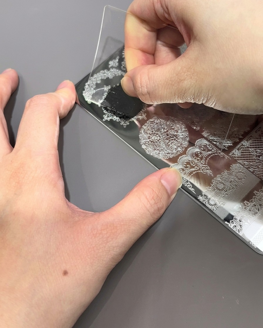 Przezroczysta plastikowa zdrapka przeciągana po metalowej płytce do stempli, usuwająca nadmiar czarnego lakieru, by odsłonić wygrawerowany wzór.