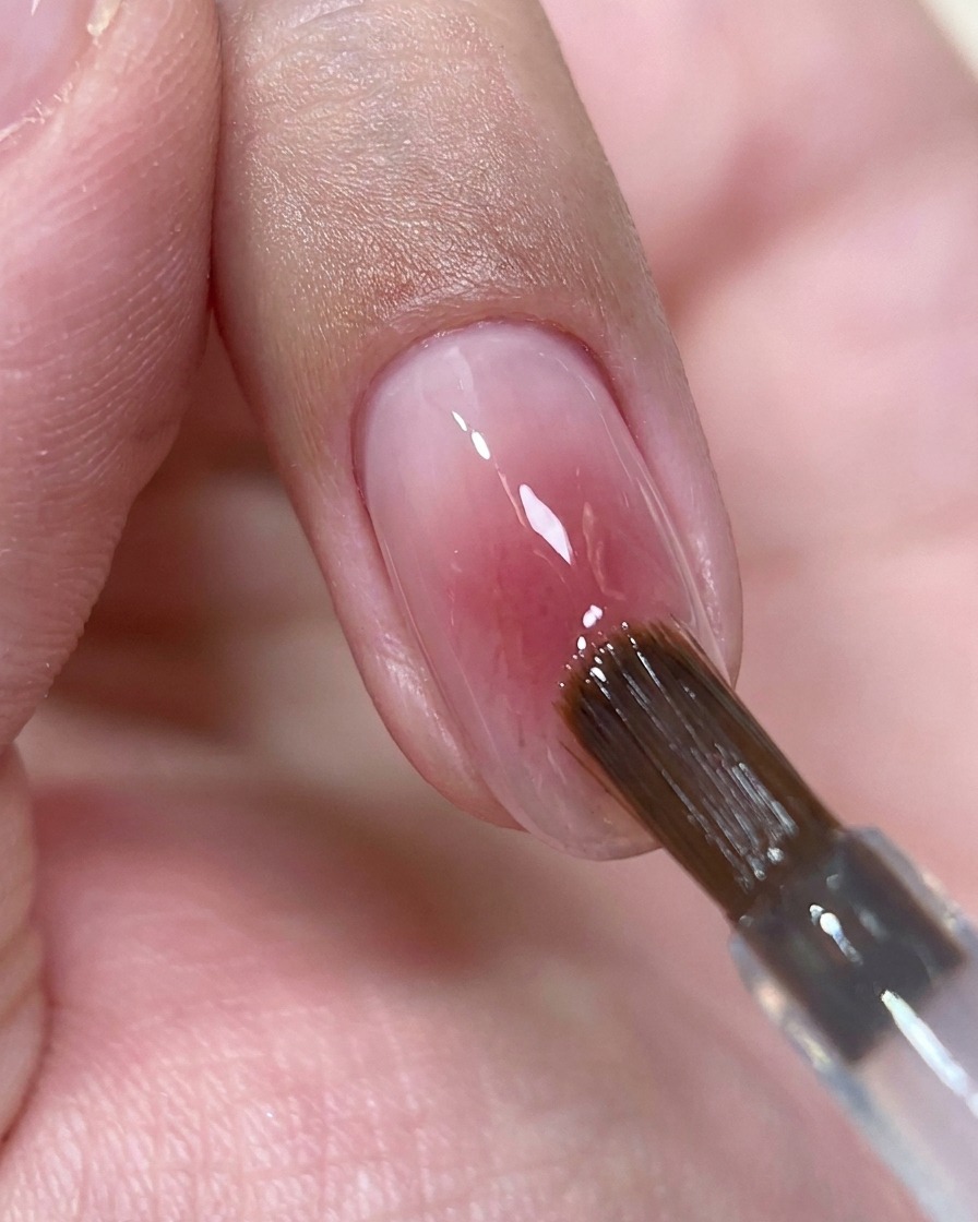 Pędzel do paznokci nakładający małą kropelkę przezroczystego żelu top coat na środek lśniącego różowego paznokcia.