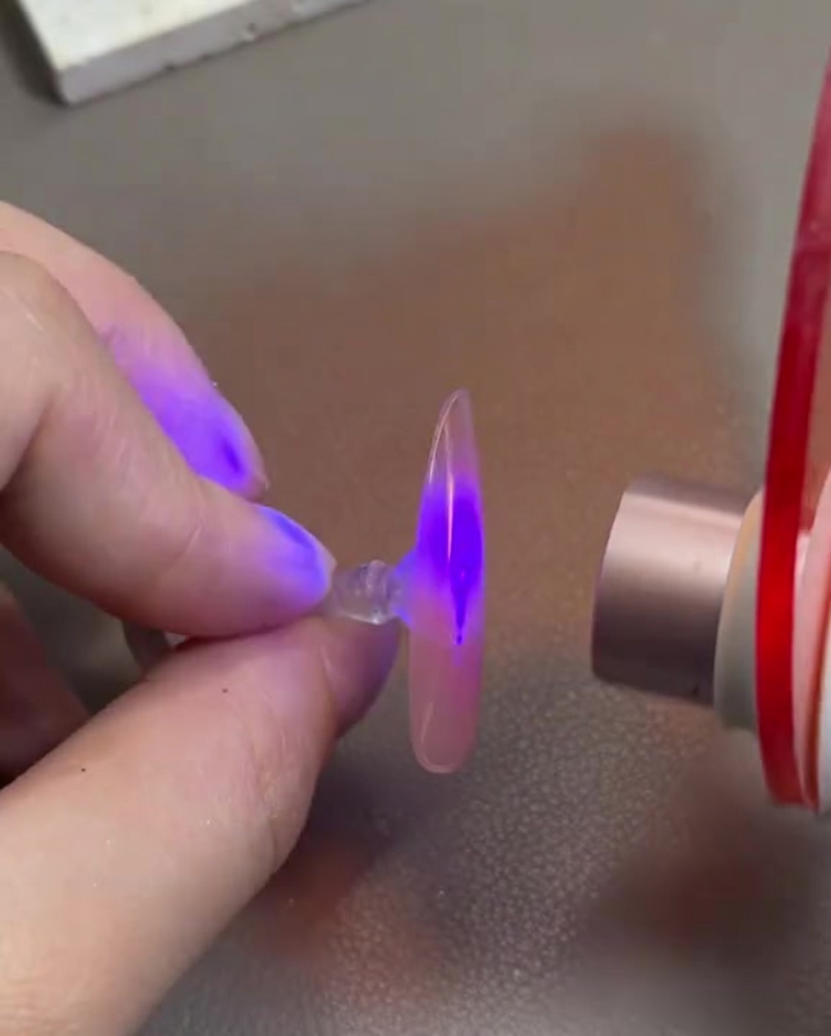 Dłoń trzymająca paznokieć z różowym żelem pod małą lampą UV/LED.