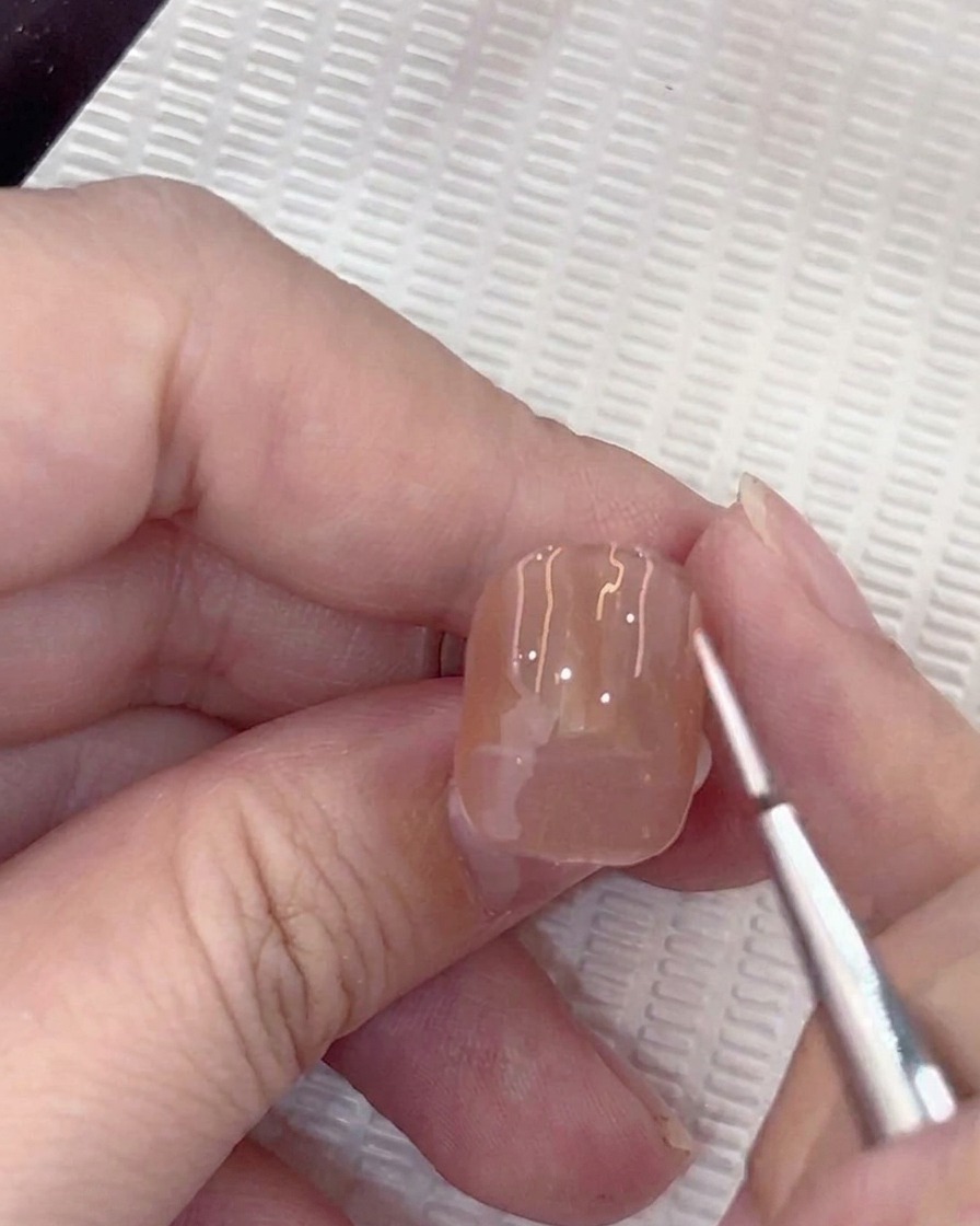 Zbliżenie na pędzelek typu liner aplikujący małą białą kropkę na środku paznokcia pomalowanego żelem w kolorze nude.