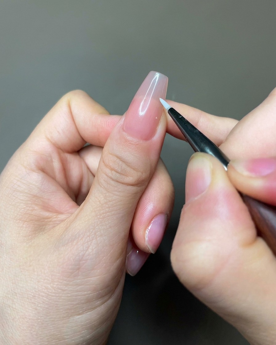 Zbliżenie na cienki pędzelek typu liner nakładający biały żel w celu stworzenia kształtów płatków na paznokciu w kolorze nude.