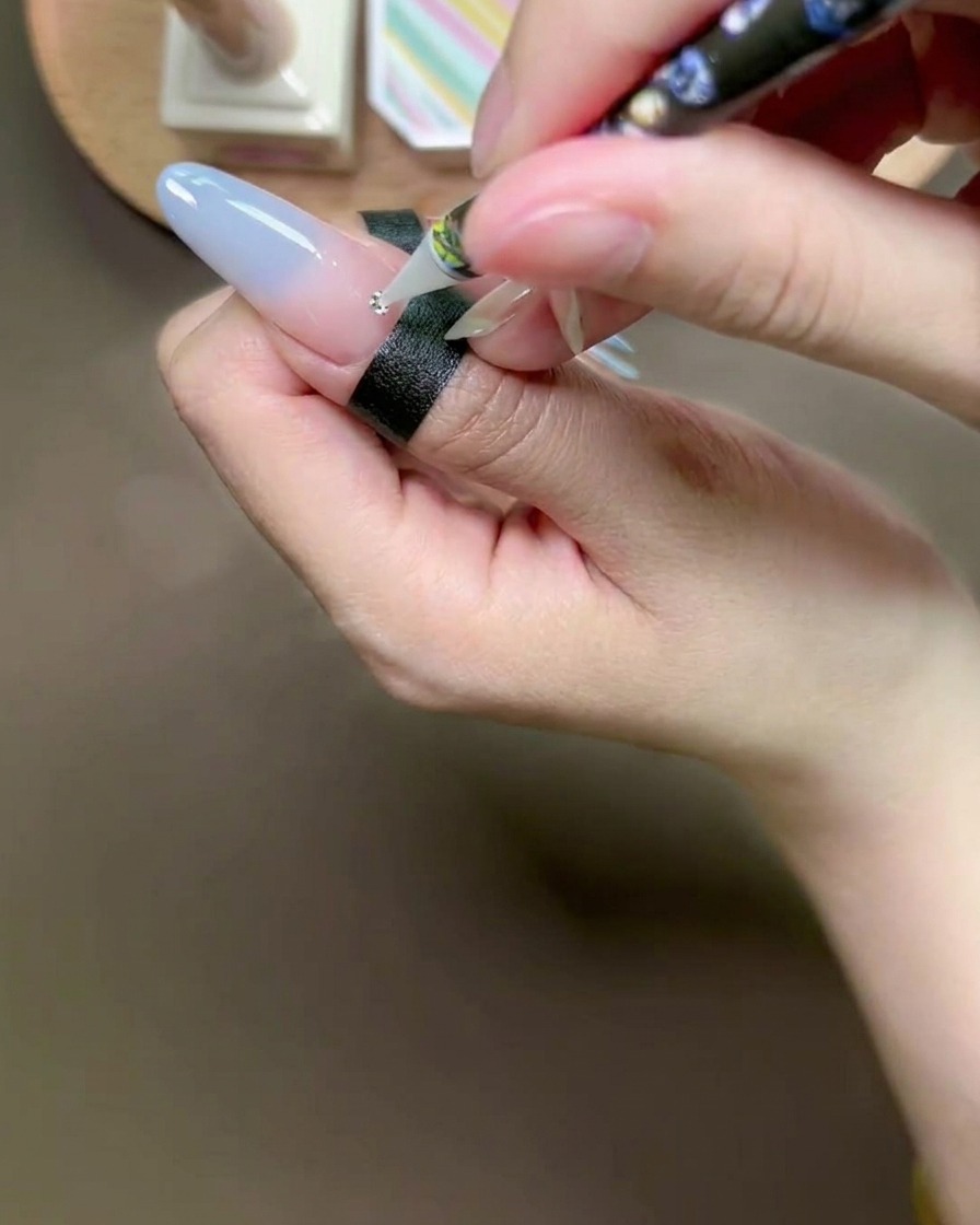 Narzędzie z woskową końcówką umieszczające małą srebrną cyrkonię na kropelce kleju na paznokciu.