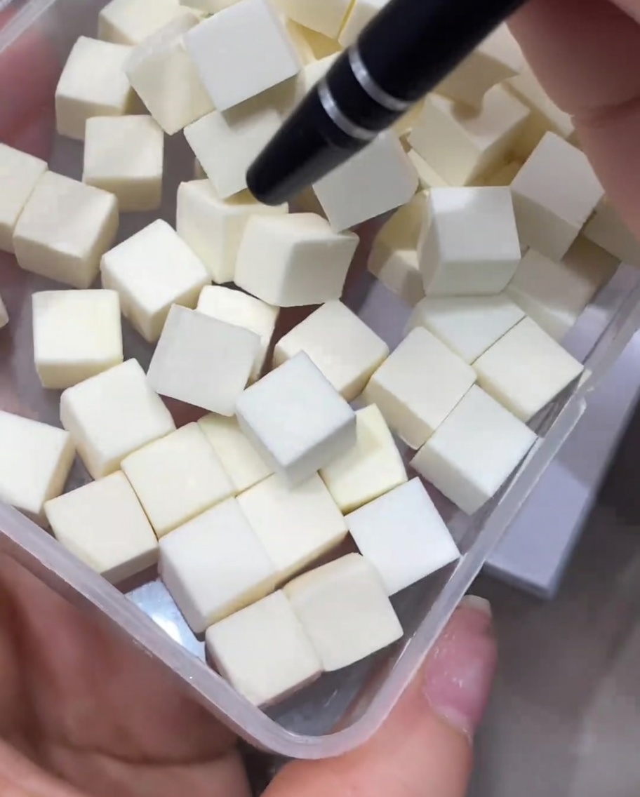 Precyzyjne narzędzie podnoszące małą, białą kostkę gąbki kosmetycznej z przezroczystego plastikowego pojemnika.