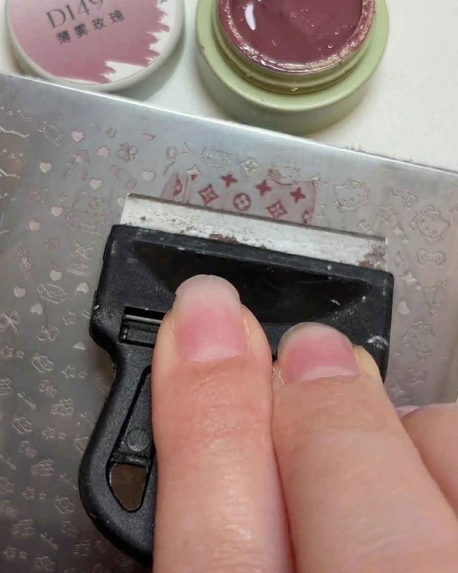 Plastikowa zdrapka rozprowadzająca niewielką ilość brudnoróżowego lakieru po grawerowanych wzorach na metalowej blaszce.
