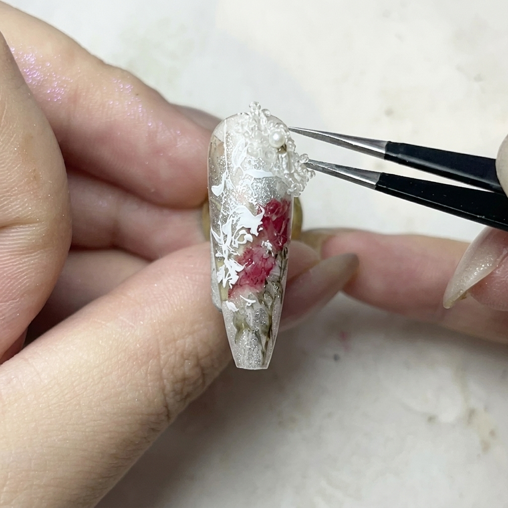 Pęseta ostrożnie umieszczająca małą białą ozdobę 3D w kształcie kwiatka na kropli żelu.