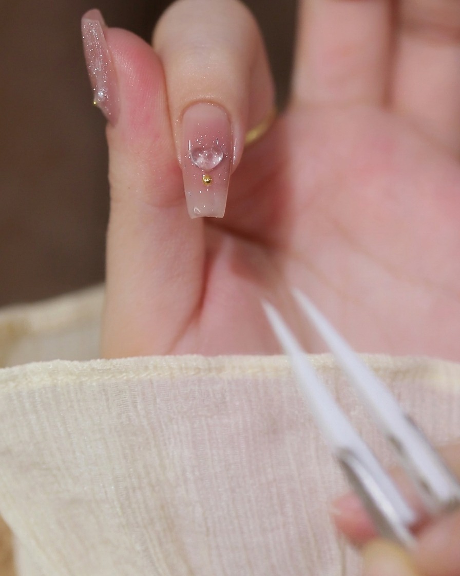 Zbliżenie na palec wskazujący z bazą mleczno-nude, z przezroczystym charms’em 3D w kształcie serca i złotym koralikiem, widoczna pęseta.