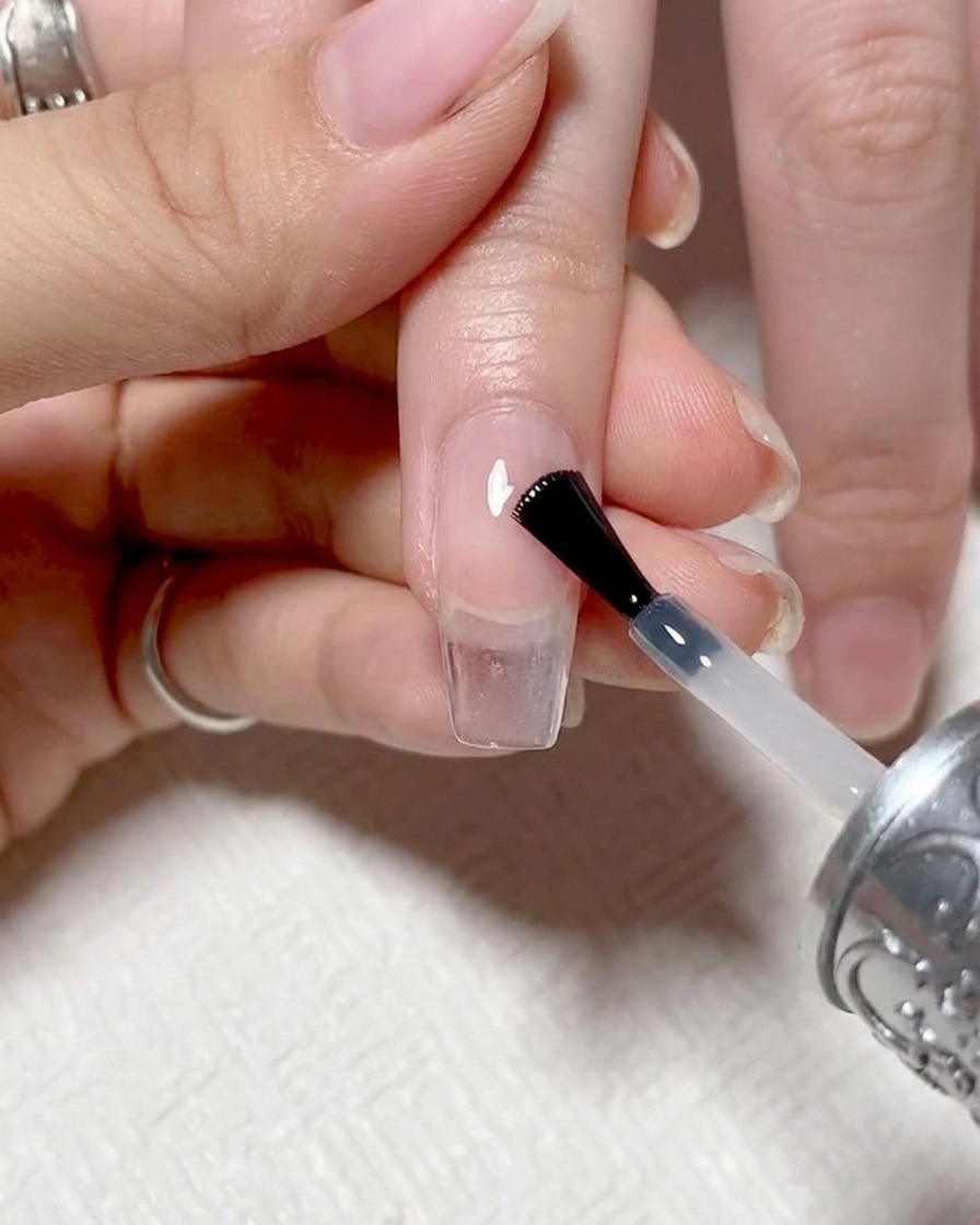 Zbliżenie pędzelka nakładającego przejrzysty, błyszczący top coat na gotowy paznokieć z gradientem.
