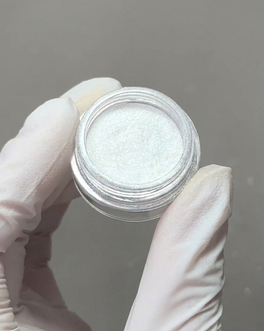 Dłoń w rękawiczce trzymająca mały pojemnik z opalizującym pyłkiem aurora z aplikatorem gąbkowym nabierającym pigment.