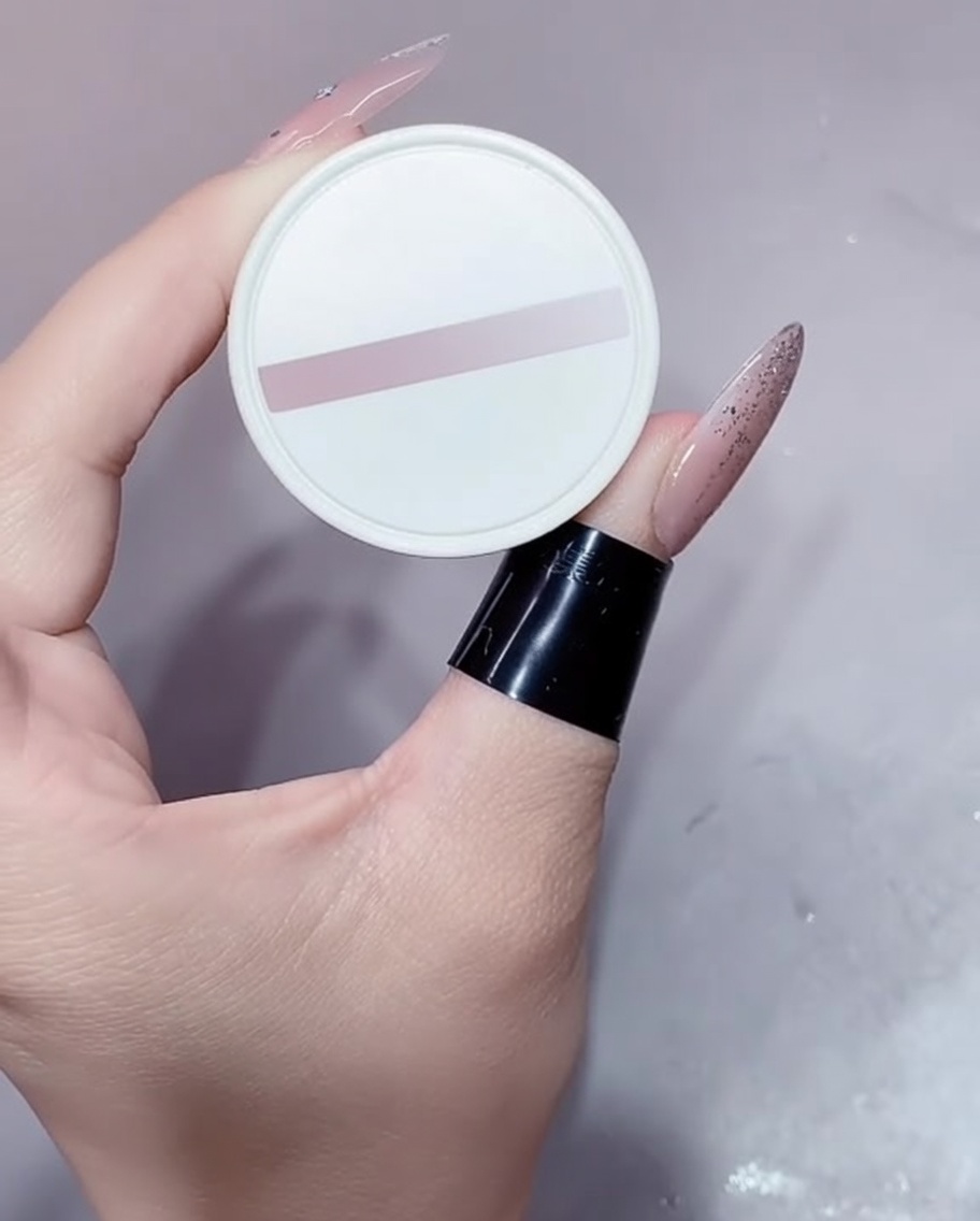Mały biały słoiczek z żelem w odcieniu nude trzymany w wypielęgnowanej dłoni.