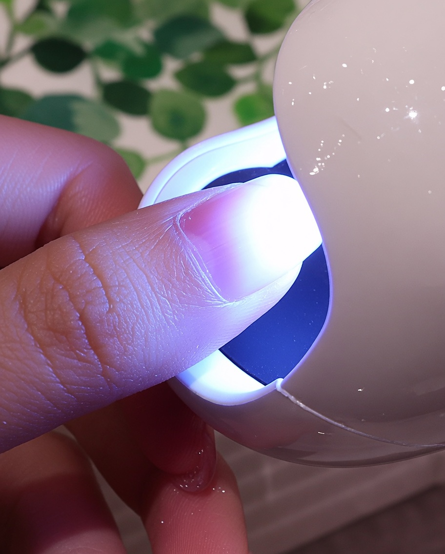 Paznokieć z teksturowaną białą powłoką umieszczony pod fioletowym światłem małej lampy UV/LED.