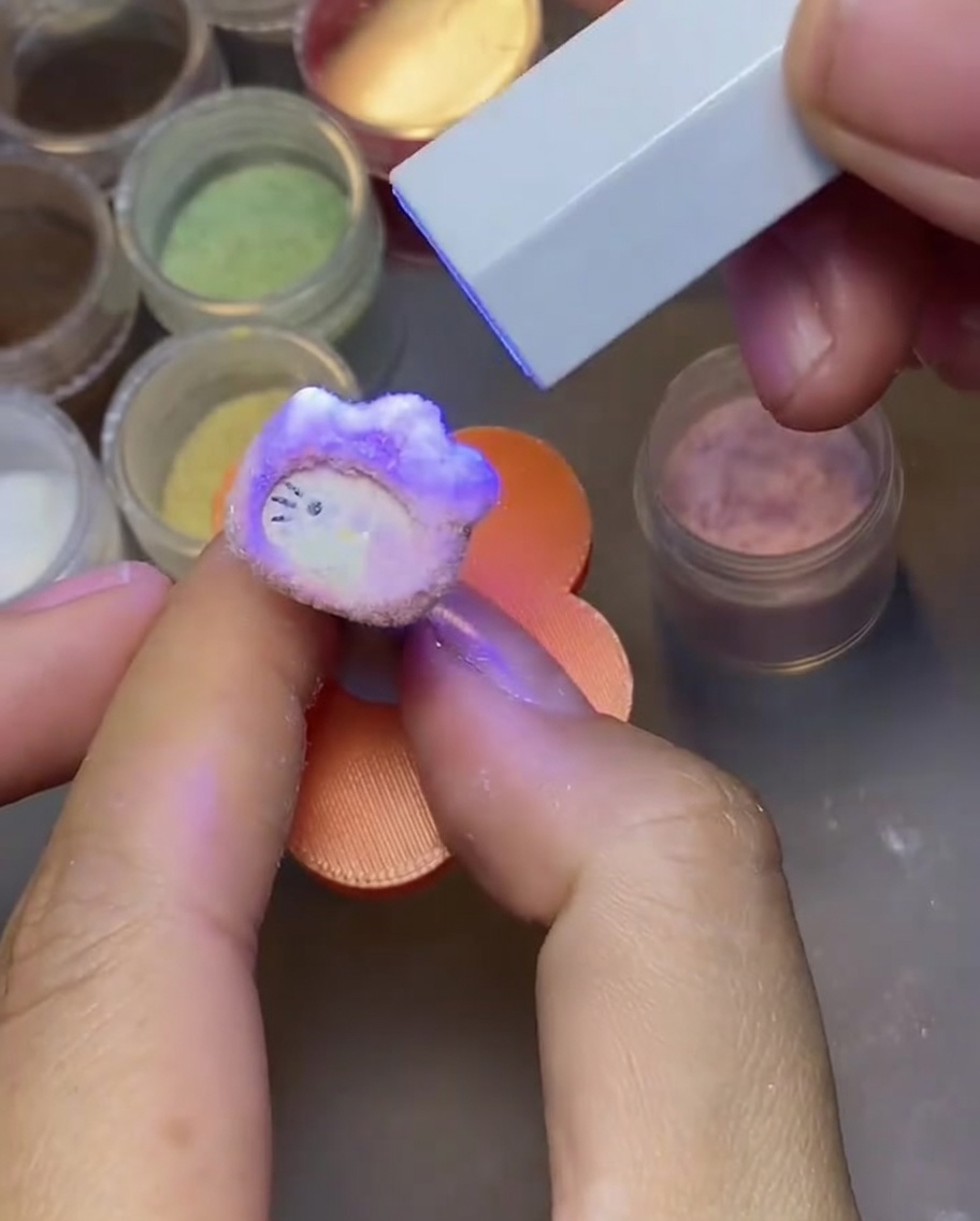 Dłoń stukająca o charms Hello Kitty po utwardzeniu, aby usunąć nadmiar pudru velvet.