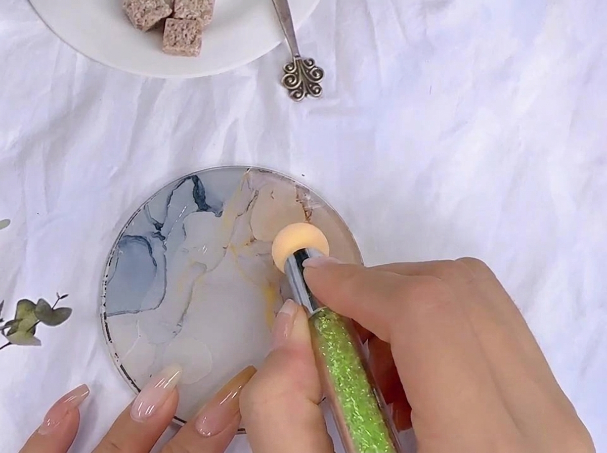 Ein Schwämmchen-Werkzeug tupft in Farbgel, das auf einer gemusterten Glas-Mischpalette verteilt ist.