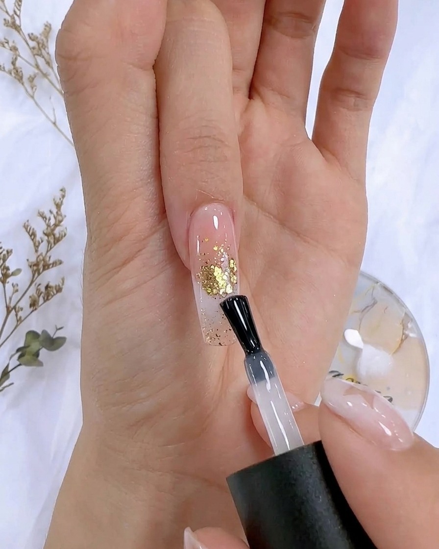 Ein Nagelpinsel trägt einen klaren Top Coat über einen dekorierten Nagel mit Blattgold-Akzenten auf.