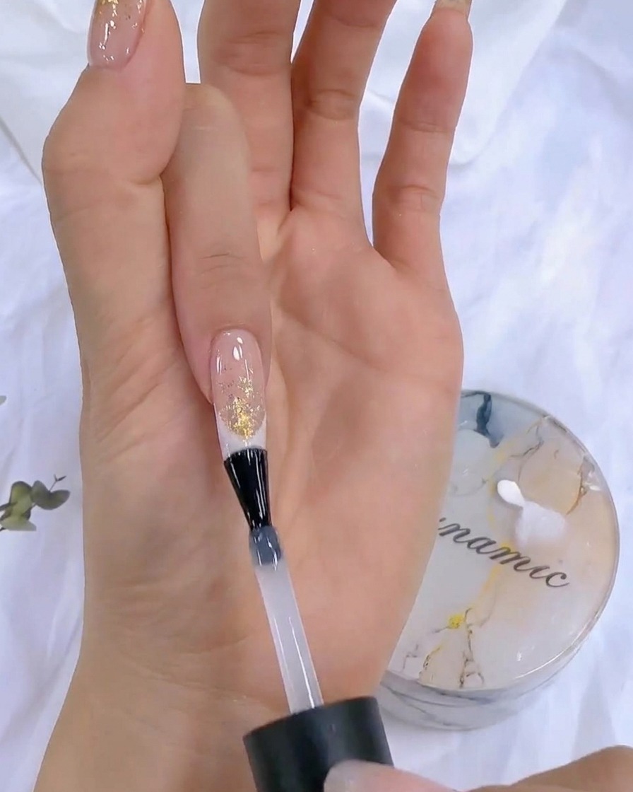 Eine Hand hält eine Top-Coat-Flasche während des finalen Aushärtungsschritts neben eine UV/LED-Nagellampe.