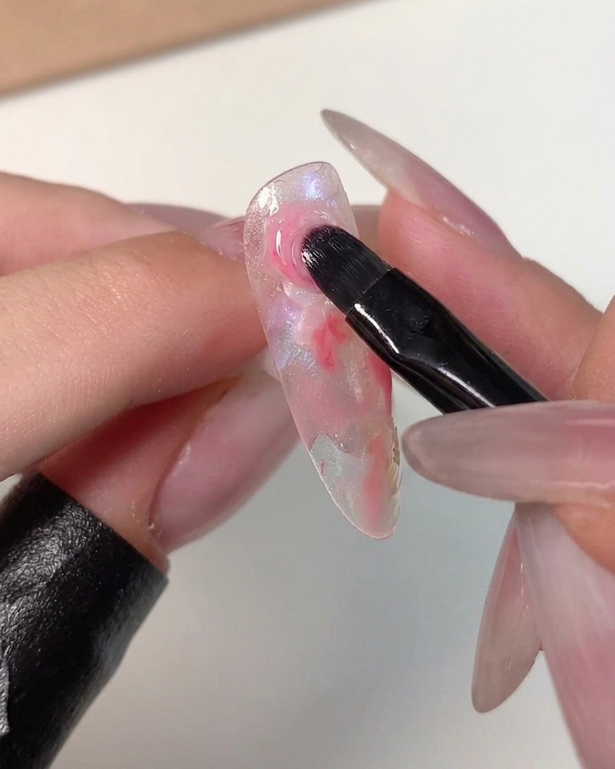 Pędzelek do nail artu rozcierający krawędzie ciemnoróżowej kropki z otaczającymi jaśniejszymi kolorami na przezroczystym tipsie.