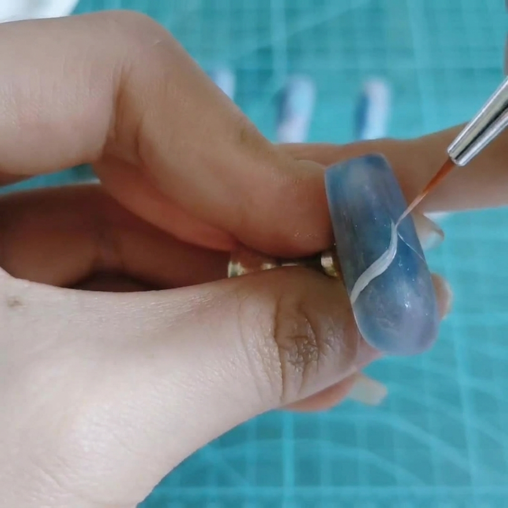 Cienki pędzelek do nail artu rysujący cienką, białą, falistą linię w poprzek niebiesko-fioletowego marmurkowego paznokcia.