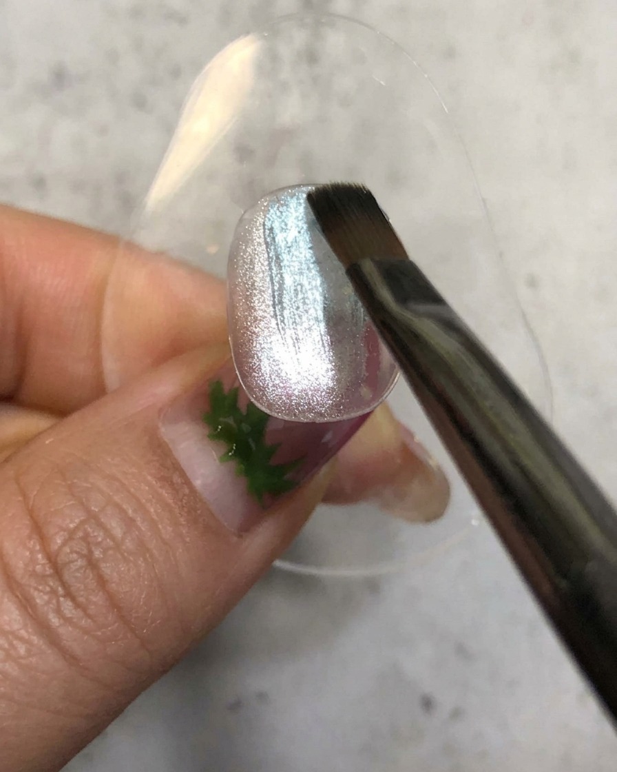 Ein Nagelpinsel trägt einen transparenten blauen Gel-Lack über einer glitzernden silbernen Magnetbasis auf.