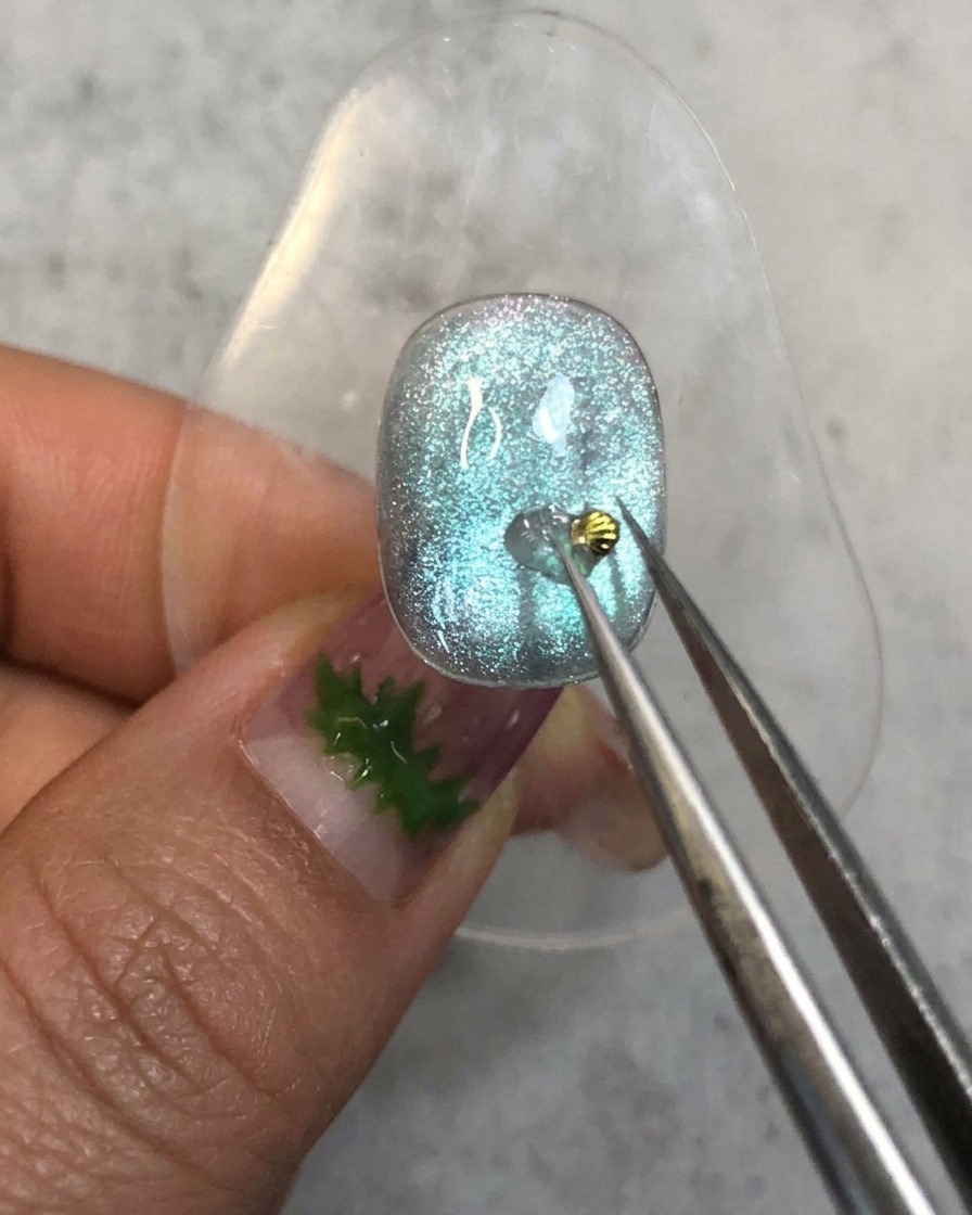 Eine Pinzette platziert einen winzigen goldenen Muschel-Charm auf den Aufbau-Gel-Punkt auf einem blauen Nagel.