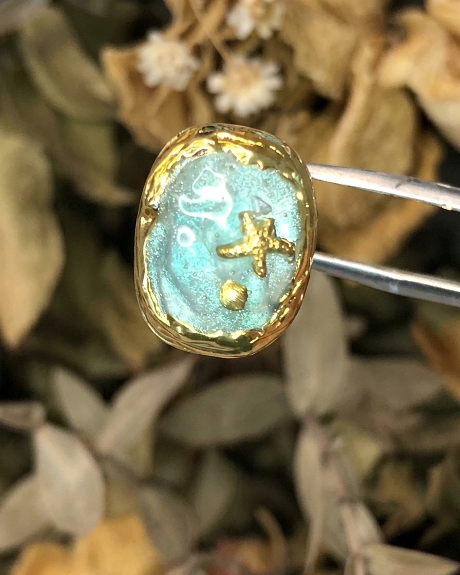 Eine Pinzette hält ein vollendetes Nageltunstwerk im Atlantis-Thema mit Goldumrahmung.