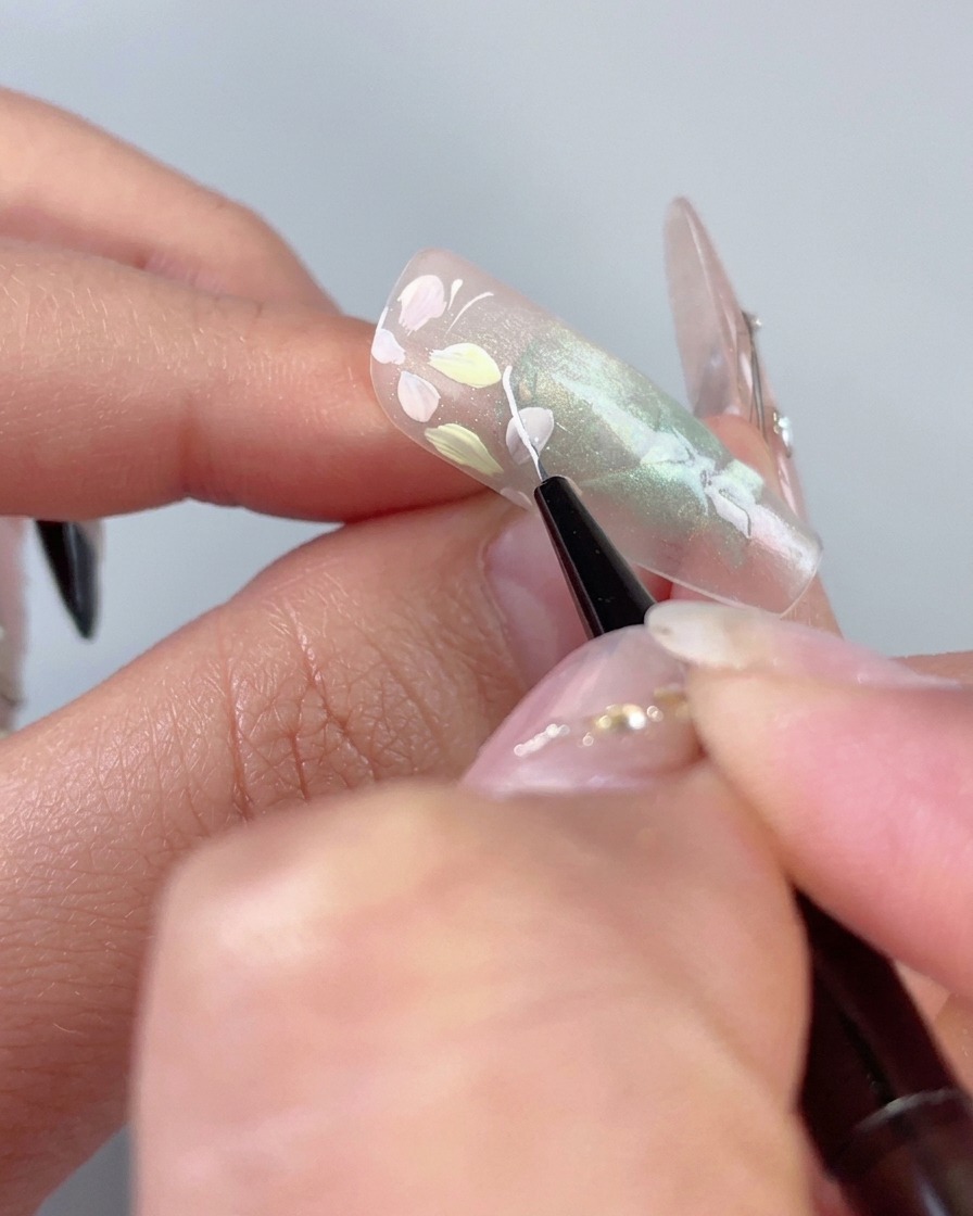 Cienki pędzel liner rysujący delikatną białą zakrzywioną linię obok pastelowych kwiatów na przezroczystym zielonym paznokciu cat-eye.