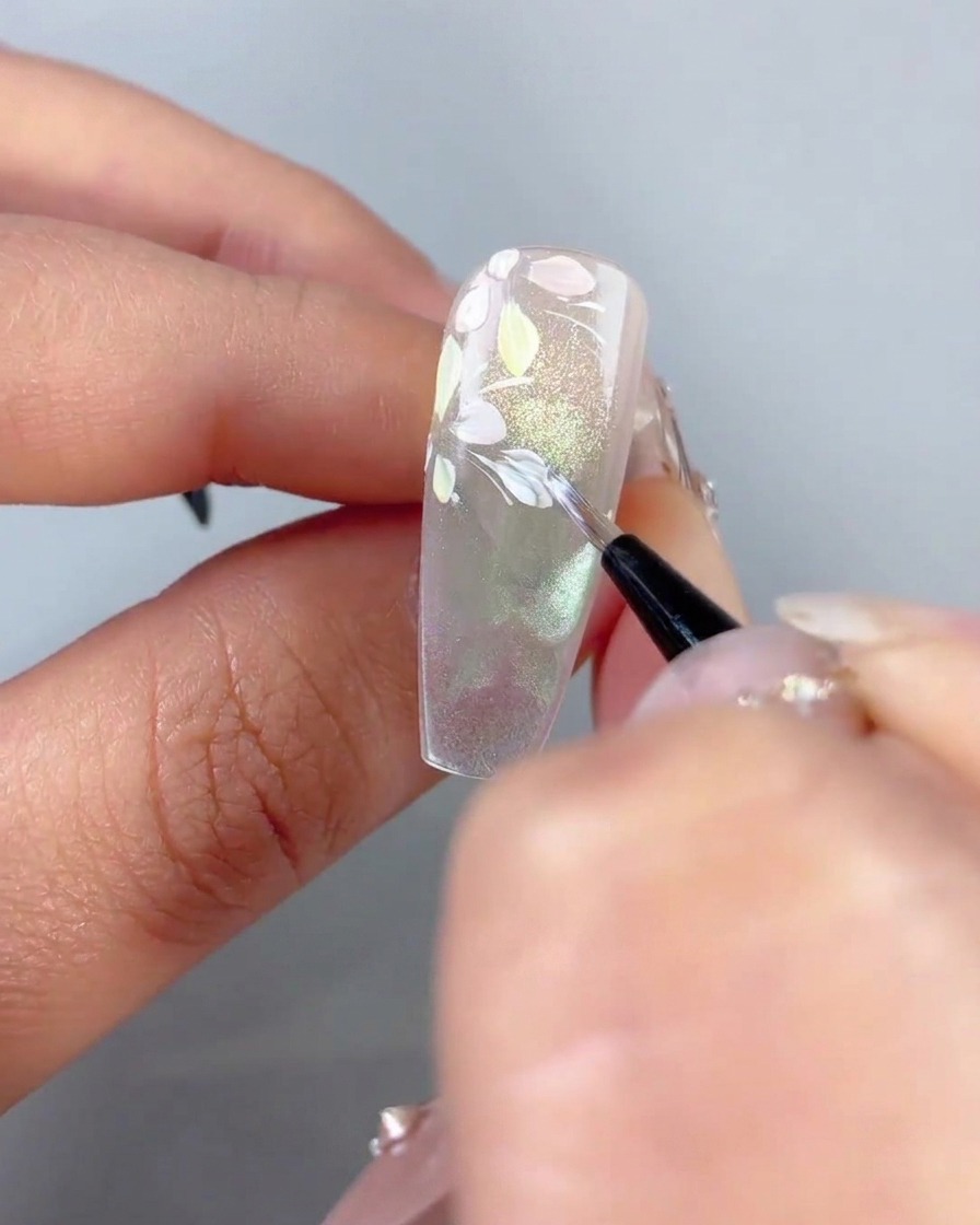 Cienki pędzel wklepujący malutkie białe akcenty liści na zakrzywionych białych liniach w pobliżu pastelowych płatków kwiatów na paznokciu.