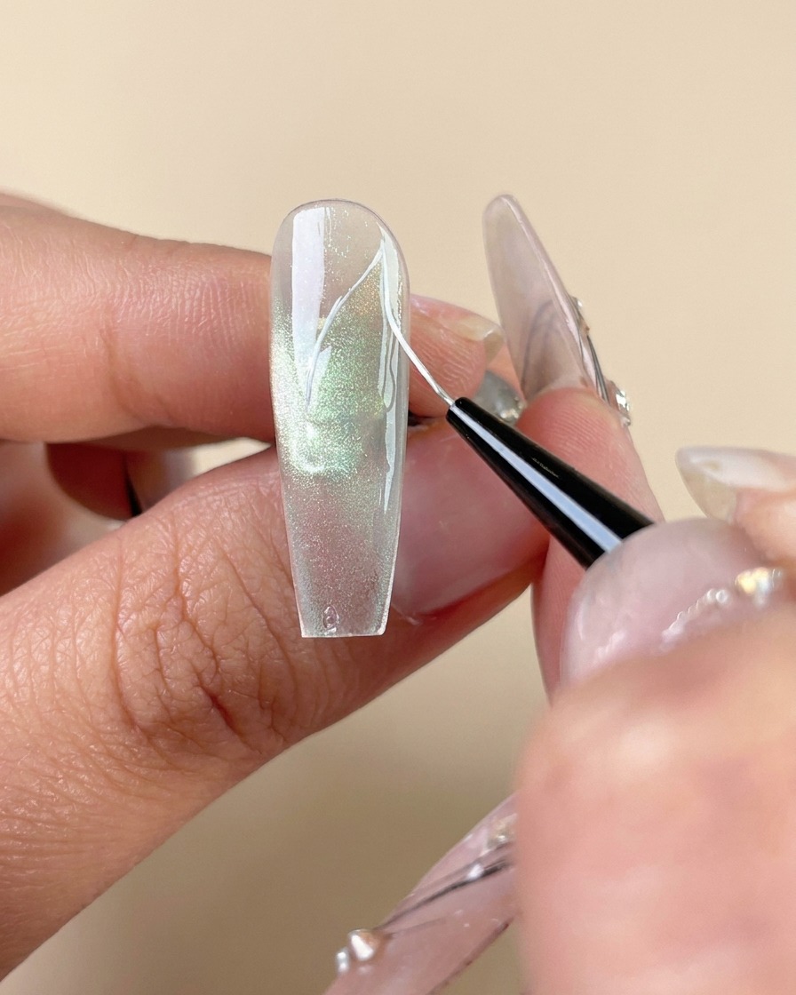 Cienki pędzel liner rysujący białe linie żyłek na zielonym paznokciu z żelem cat-eye.