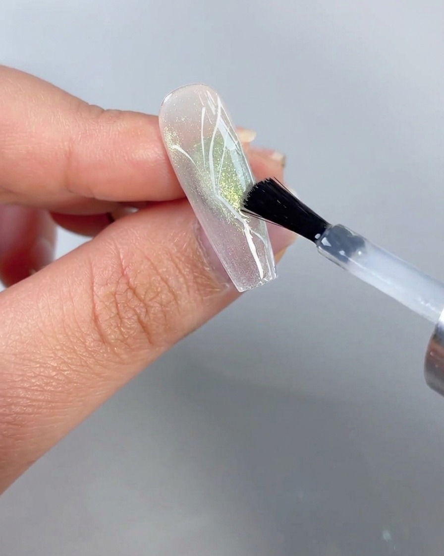 Pędzel nakładający przezroczysty top coat na paznokieć z namalowanym projektem półskrzydła motyla na tle przezroczystego zielonego cat-eye.