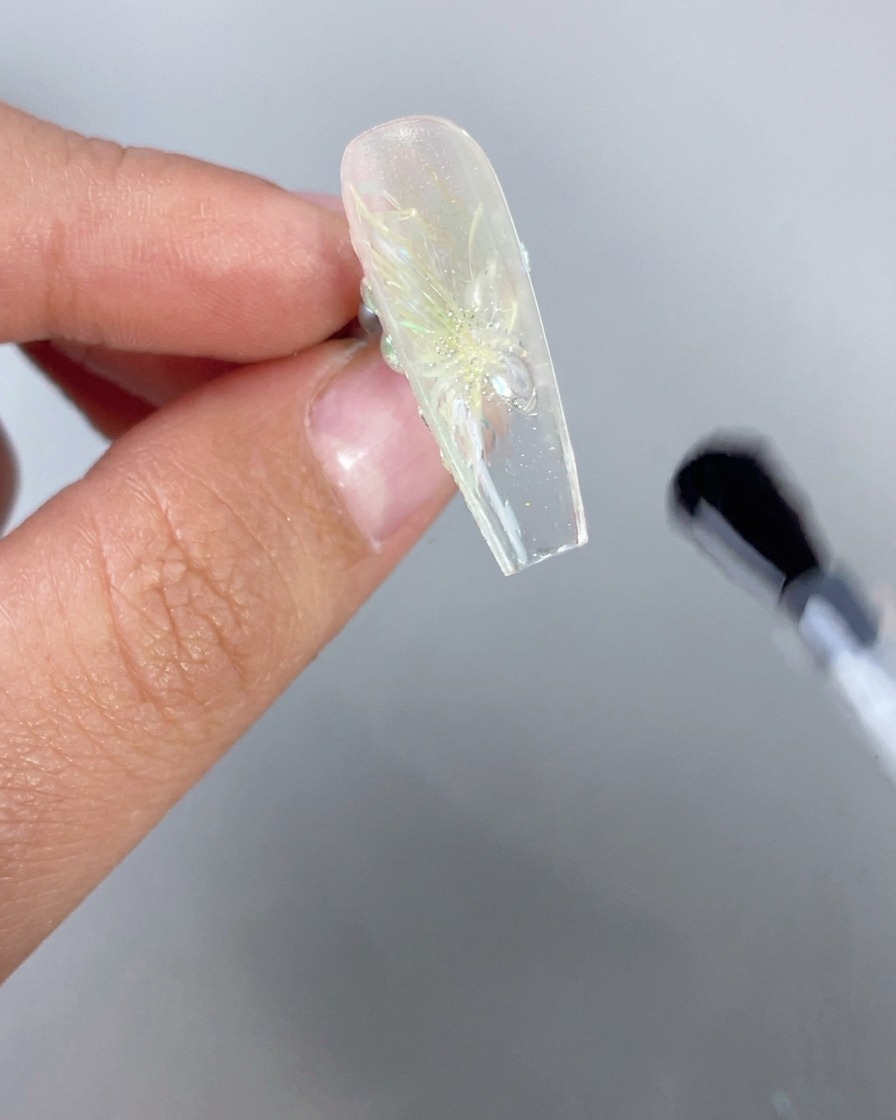 Zbliżenie dłoni trzymającej przezroczysty lód paznokieć z motywem kwiatowym 3D do góry dnem, podczas gdy pędzel nakłada przezroczysty top coat na spód wolnego brzegu.