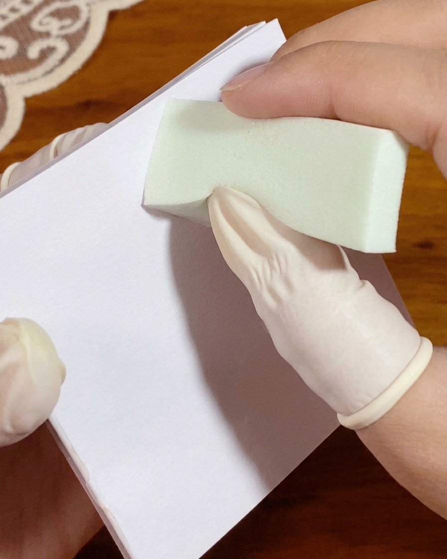 Ręka w rękawiczce dociskająca gąbkę z białym żelem do papieru paletowego przed aplikacją na paznokieć.