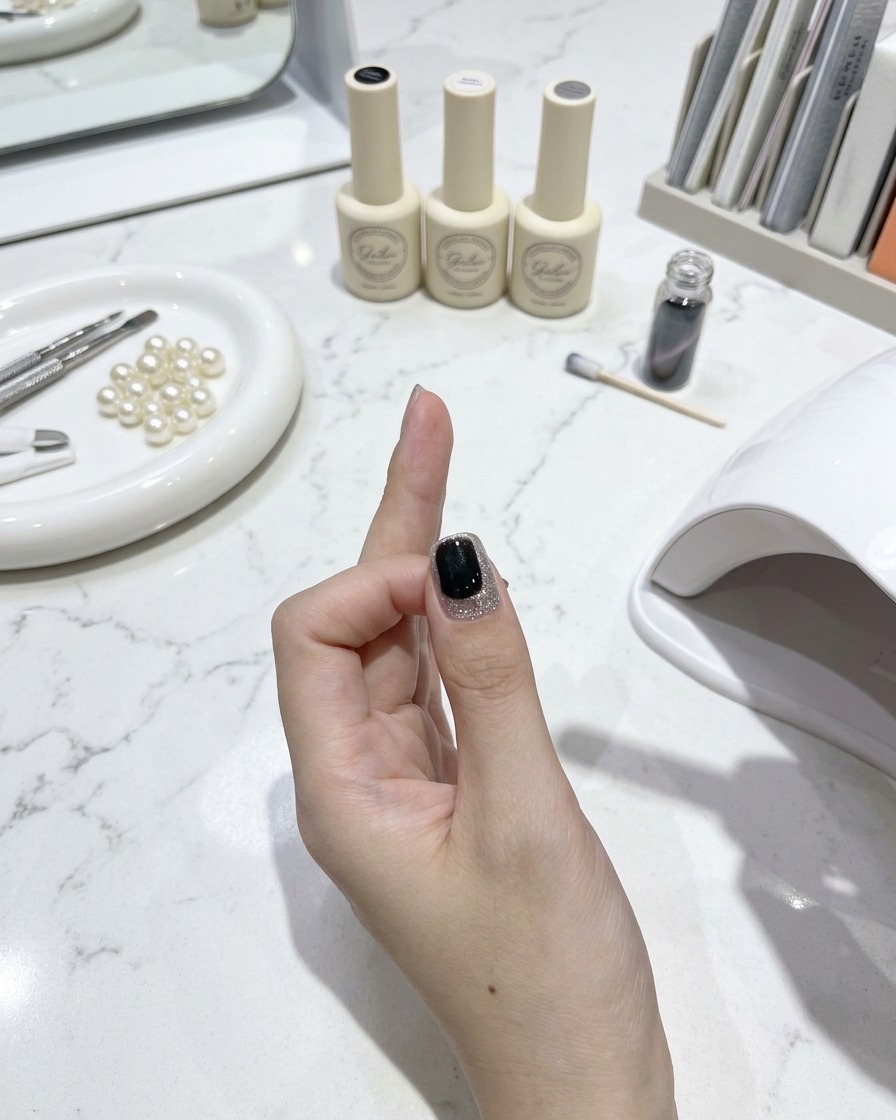 Pędzelek do paznokci nakładający błyszczący top coat na gotowy paznokieć cat-eye w otoczeniu przyborów do manicure i buteleczek z lakierem na białym marmurowym stole w tle.