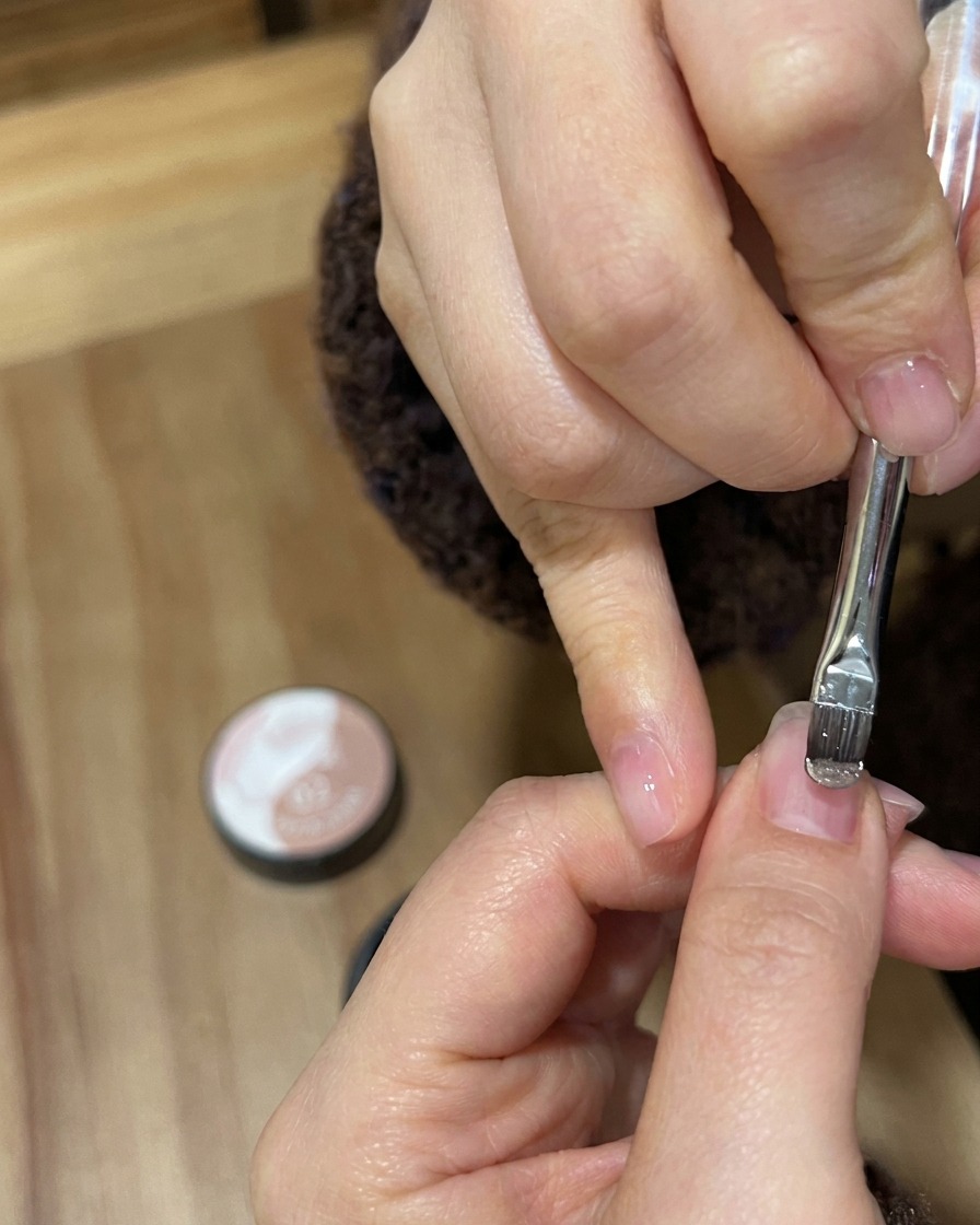 Zbliżenie pędzelka do żelu nakładającego mieniący się lakier nude gold na naturalną płytkę paznokcia.