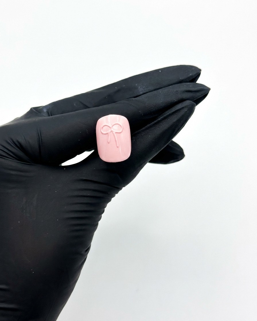 Ręka w rękawiczce trzymająca różowy sztuczny paznokieć pod lampą UV do paznokci, aby utwardzić wzór kokardki 3D.