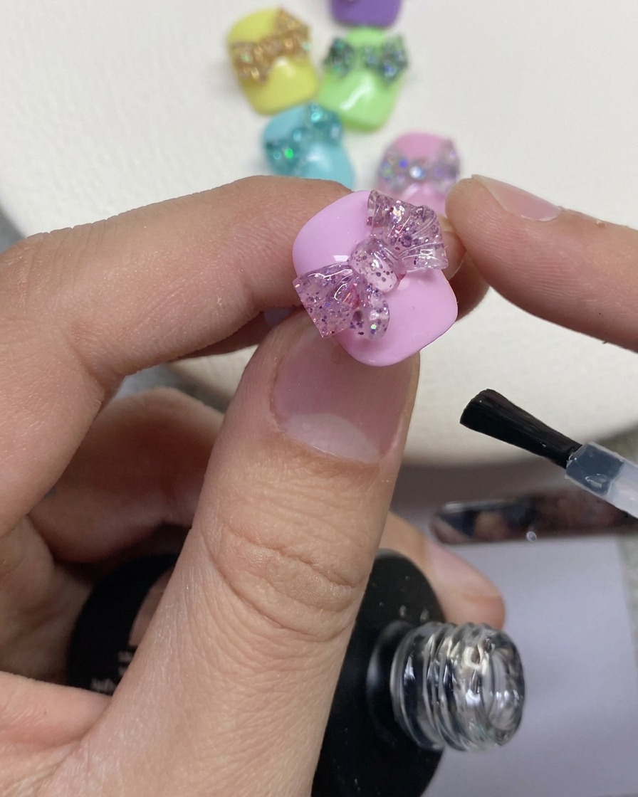 Zbliżenie na pędzelek nakładający przezroczysty top coat wokół brokatowej kokardy 3D na pomalowanym różowym paznokciu.