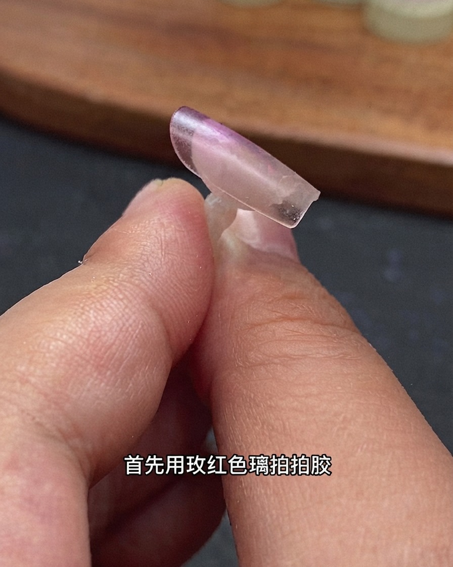 Eine Nahaufnahme einer klaren Nagelverlängerungsspitze, die von Fingern gehalten wird, mit rosarotem Glasgel am hinteren Rand.