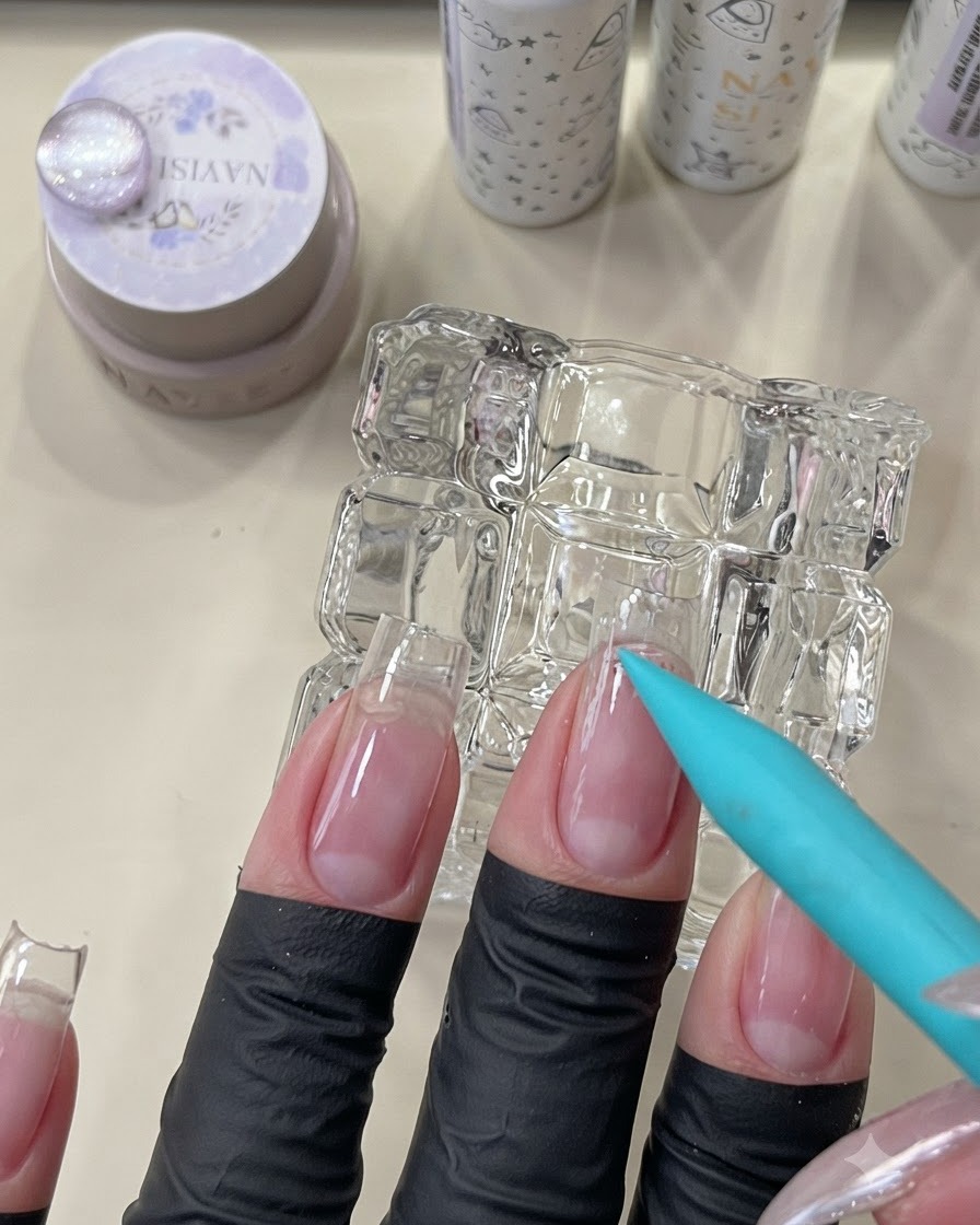 Niebieski pędzelek nakładający bezbarwną bazę peel-off w okolicy skórek naturalnego paznokcia.