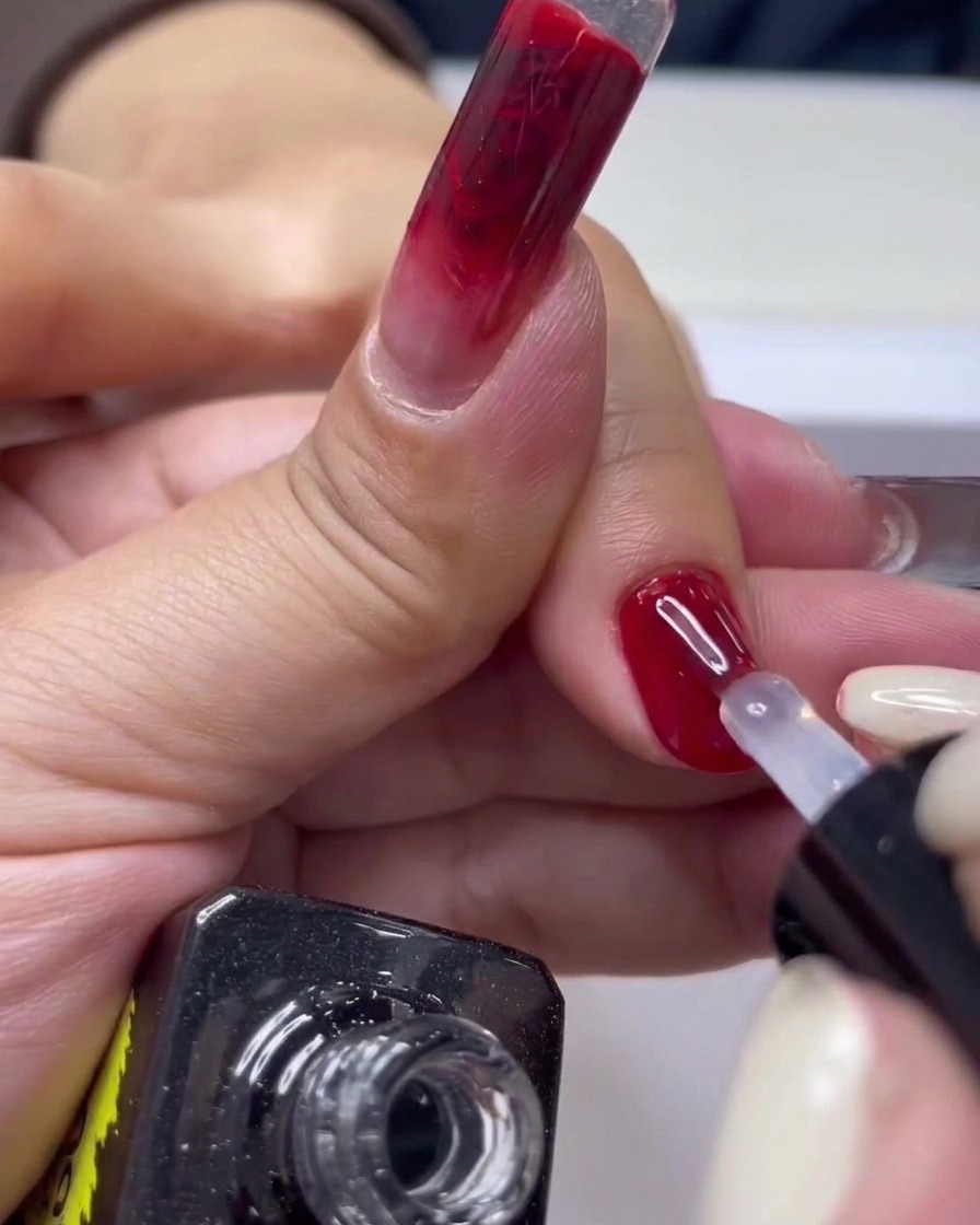 Zbliżenie pędzelka do paznokci nakładającego nawierzchniowy top coat na błyszczący czerwony manicure żelowy.