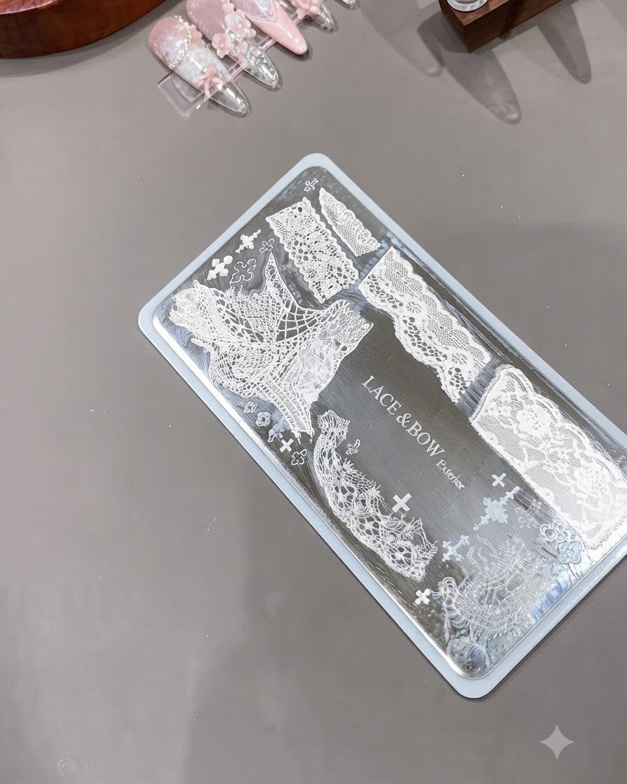 Przezroczysty silikonowy stempel do paznokci umieszczony obok grawerowanej metalowej płytki przedstawiającej skomplikowane wzory białej koronki.