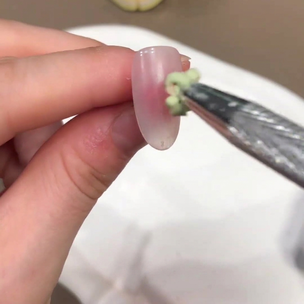 Ein kleiner Pinsel trägt einen grün getönten Geltropfen auf die Mitte eines nudefarbenen Nagels auf.