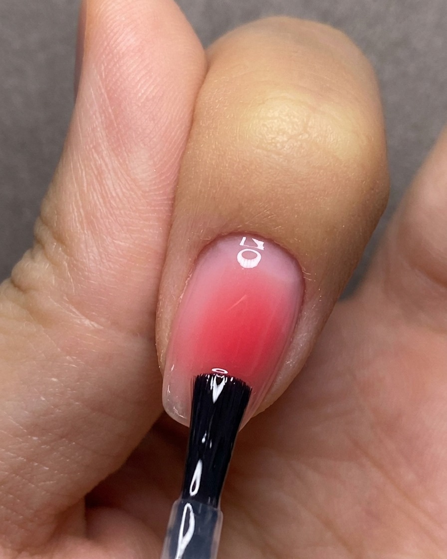 Pędzelek równomiernie nakładający bezbarwny top coat na paznokieć z czerwonym gradientem blush.