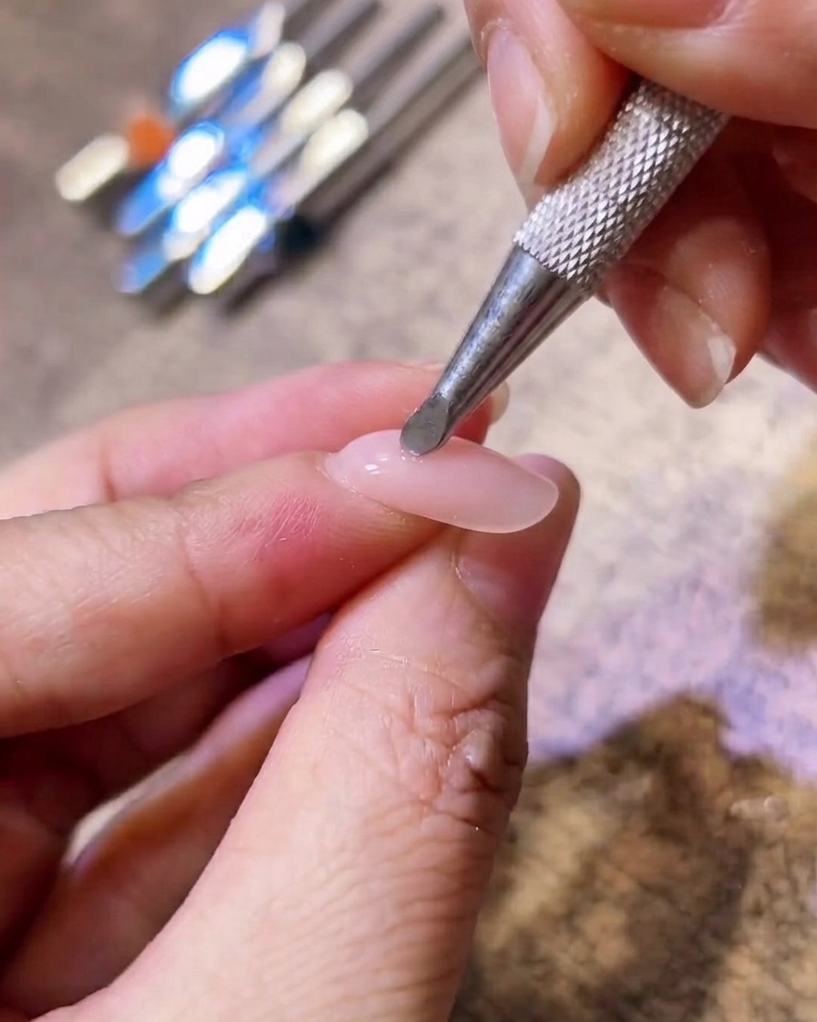 Zbliżenie na metalowy aplikator nakładający różowy pigment na środek paznokcia żelowego w kolorze nude.