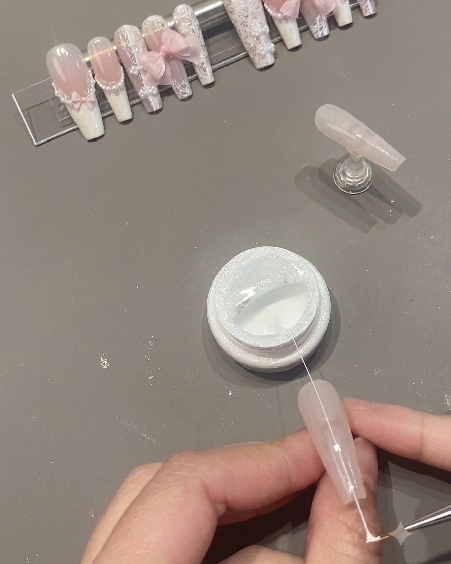 Wąskie pasmo białego żelu spider gel przeciągane nad powierzchnią paznokcia obok otwartego słoiczka.