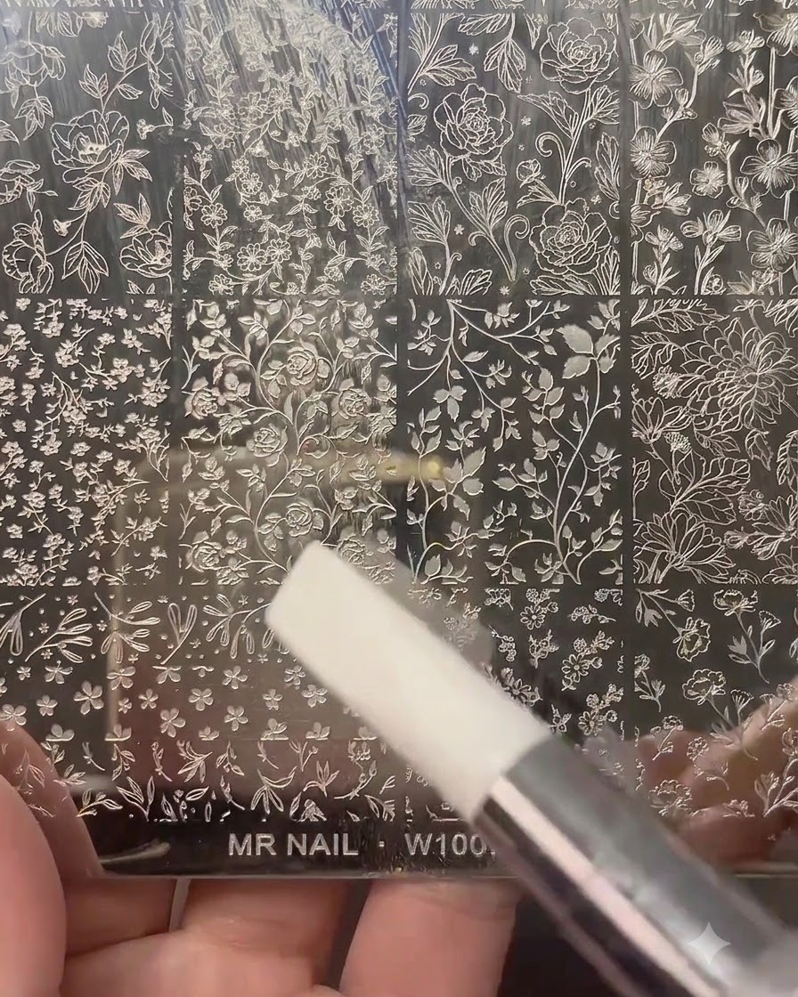 Przezroczysty stempel do paznokci naciskający na wygrawerowaną metalową blaszkę do stempli wypełnioną motywami kwiatowymi.